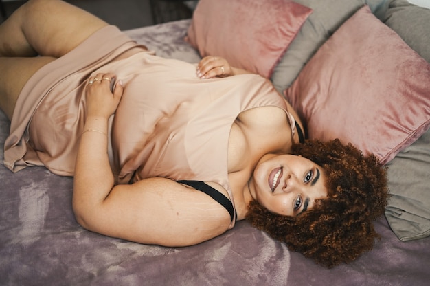 Piękna krągła plus size afrykańska czarna kobieta afro włosy leżące na łóżku w jedwabnej pudrowo różowej sukience przytulnej