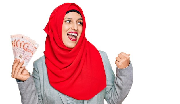 Piękna latynoska kobieta ubrana w islamski hidżab trzymająca banknoty dolarów singapurskich wskazująca kciukiem na bok uśmiechnięta zadowolona z otwartymi ustami