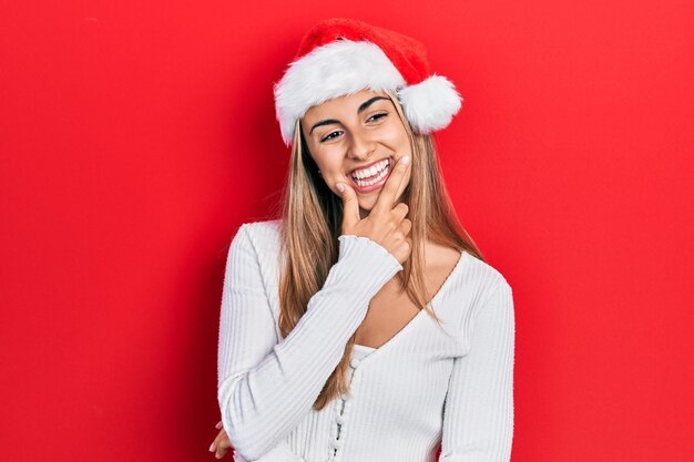 Piękna latynoska kobieta w świątecznym kapeluszu, wyglądająca pewnie przed kamerą, uśmiechnięta ze skrzyżowanymi ramionami i ręką podniesioną na brodzie, myśląca pozytywnie