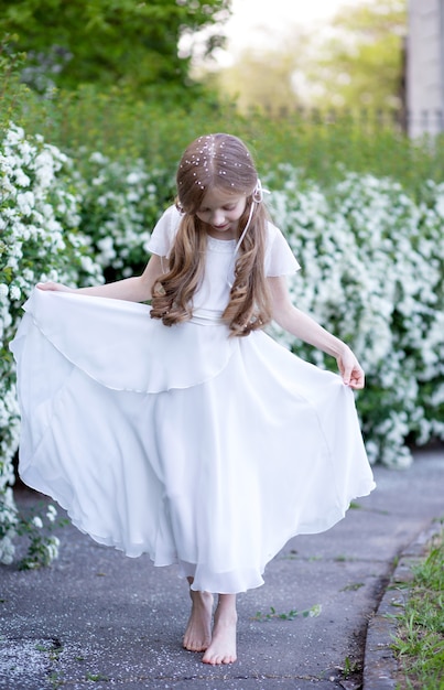 piękna mała blondynka w wieku 9 lat, z długimi włosami w białej jedwabnej sukience, tańcząca balet w parku, spoglądająca w dół, jej głowa ma wiele małych białych płatków