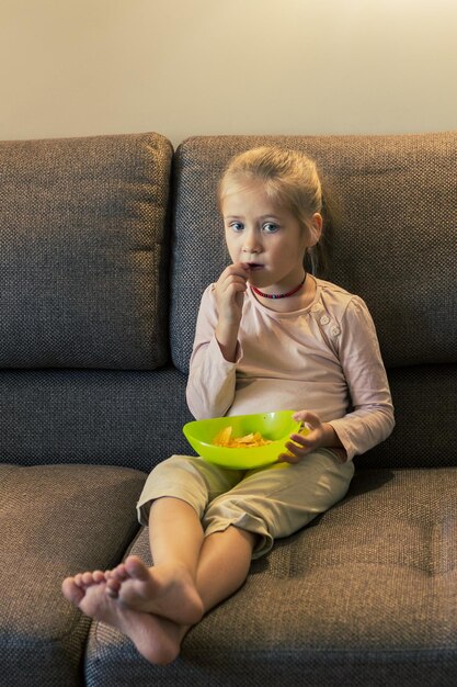 Piękna mała dziewczynka je niezdrowe jedzenie podczas oglądania telewizji na kanapie