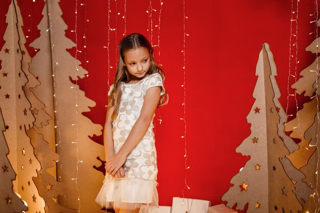 Piękna mała dziewczynka marzy o prezentach świątecznych. Czerwone ozdoby świąteczne, niezwykłe. Wieczór przed Bożym Narodzeniem, Wigilia