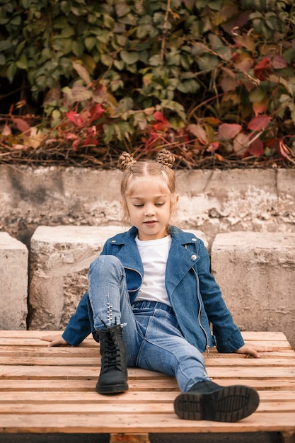 Piękna mała dziewczynka siedzi w stylowych ubraniach na drewnianych paletach. Stylowe dziecko w niebieskiej kurtce. Styl casual, moda dla dzieci, modny garnitur, szczęśliwe dzieciństwo!