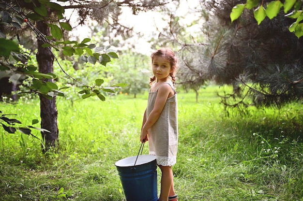 Piękna mała dziewczynka w lnianej sukience stoi w ogrodzie z dużym niebieskim metalowym wiadrem w dłoniach o zachodzie słońca