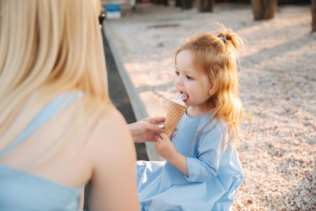 Piękna mała dziewczynka w niebieskiej sukience jedząca lody Mama pomaga i wyciera usta