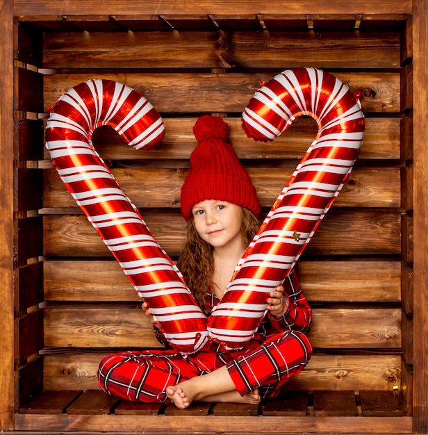 Piękna mała dziewczynka w świątecznej piżamie trzyma w dłoni duże karmelowe cukierki na drewnianym tle