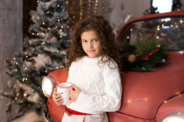Piękna mała dziewczynka z kubkiem świątecznym w pobliżu czerwonego samochodu i choinek ze światłami. Wesołych Świąt i Wesołych Świąt