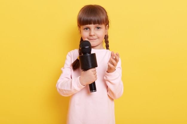 Piękna Mała Dziewczynka Z Mikrofonem, Czarujący Uśmiech, Podnosząca Rękę, Wygląda Na Nieco Nieśmiałego, Pozuje Model Dziecka Na Białym Tle Na żółtej ścianie.