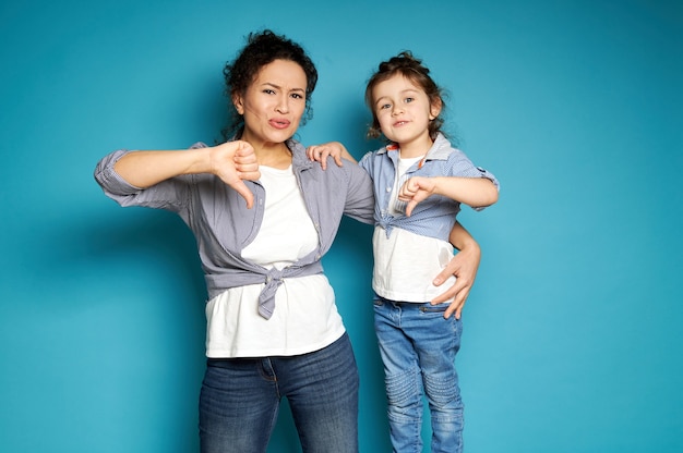 Piękna mama i córka jednakowo ubrane, pokazujące kciuki w dół i wyrażające niezadowolenie i niezadowolenie, pozując przodem na niebieskiej powierzchni z miejscem na kopię