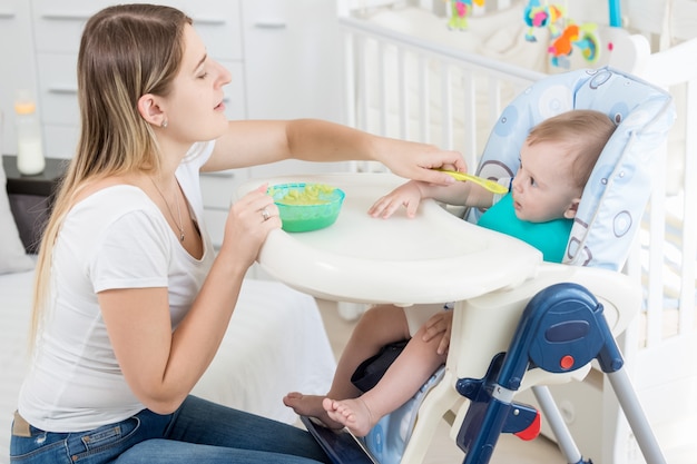 Piękna mama karmi swojego chłopca w krzesełku z owsianką