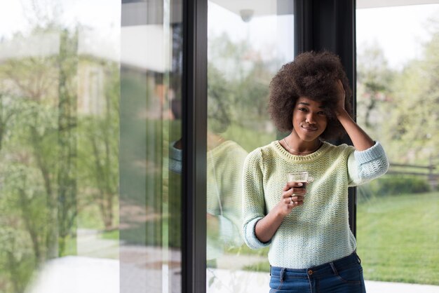 Piękna młoda afroamerykanka pijąca kawę i patrząca przez okno w swoim luksusowym domu