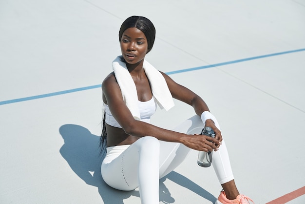 Piękna młoda afrykańska kobieta w sportowej odzieży trzymająca butelkę z wodą siedząc na sportowym t...