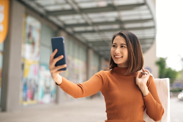 Piękna młoda Azjatka trzymająca telefon do selfie z torbą na zakupy w centrum handlowym na świeżym powietrzu