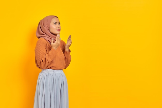 Piękna młoda Azjatka w brązowym swetrze i hidżabie, ręce złożone w geście modlitwy, błagając o coś na białym tle na żółtym tle. Koncepcja religijna ludzi islamu