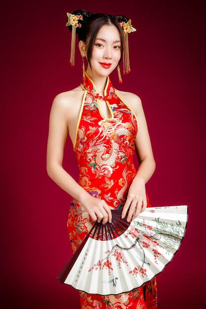 Piękna młoda Azjatka z czystą, świeżą skórą, ubrana w tradycyjną sukienkę cheongsam qipao, trzymająca wentylator pozowanie na czerwonym tle Portret modelki w studio Szczęśliwego chińskiego nowego roku