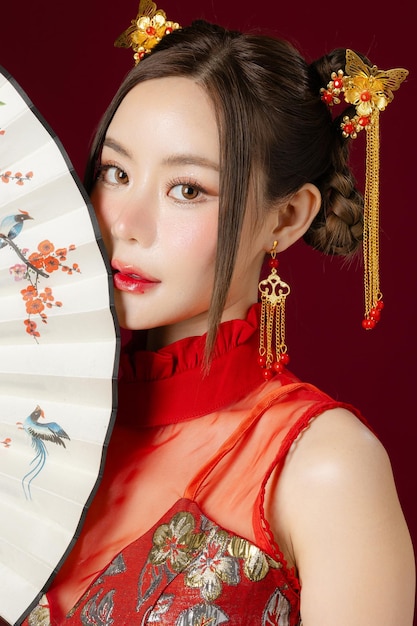 Piękna młoda Azjatka z czystą, świeżą skórą, ubrana w tradycyjną sukienkę cheongsam qipao, trzymająca wentylator pozowanie na czerwonym tle Portret modelki w studio Szczęśliwego chińskiego nowego roku