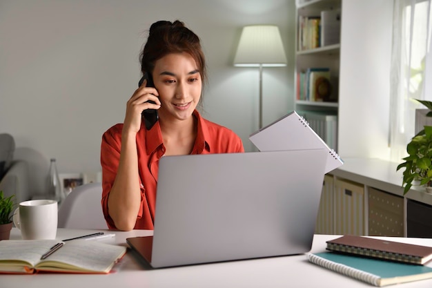 Piękna młoda Azjatycka kobieta pracuje w domu z laptopem i rozmawia na smartfonie i uśmiecha się. Kobieta pracuje na komputerze i korzysta z telefonu komórkowego siedząc w domu
