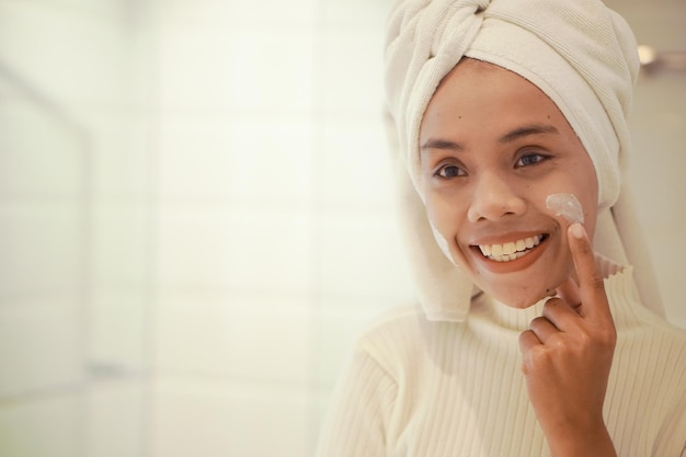 Piękna młoda azjatycka kobieta w białej szacie nakładająca balsam na twarz w łazience przed dużym lustrem