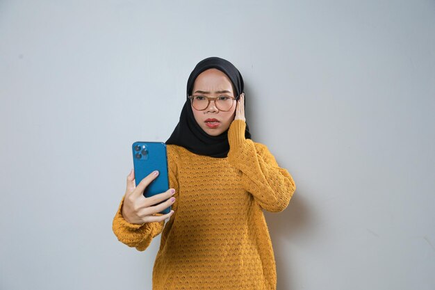 Piękna młoda azjatycka muzułmanka ubrana w pomarańczowy sweter i okulary podczas korzystania z telefonu komórkowego