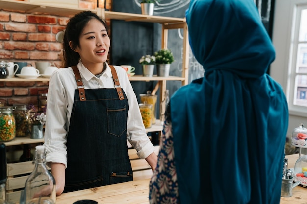 piękna młoda azjatycka restauracja kobieta właściciel w fartuch rozmawia z muzułmańskim klientem w niebieskim hidżabie przy kasie w porannej kawiarni. pani kelnerka przyjmowanie zamówienia z malajski kobieta w nowoczesnej kawiarni.