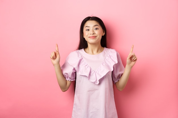Piękna młoda azjatykcia kobieta w romantycznej sukience, wskazując palcami w górę, uśmiechając się i chętnie pokazując reklamę, stojąc na różowym tle.