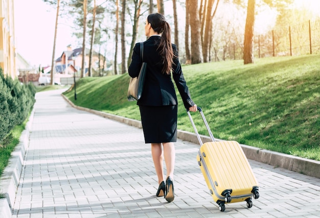 Piękna młoda biznesowa kobieta w garniturze i butach spacerująca po ulicy miasta z żółtą walizką podróżną