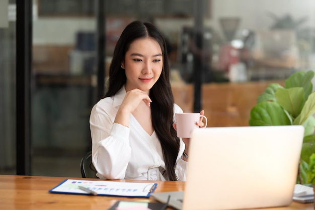 Piękna młoda bizneswoman azjatyckich picia kawy pracy na laptopie w biurze.