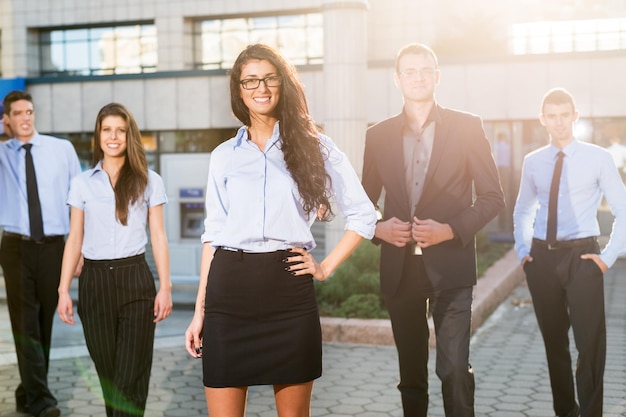 Piękna młoda bizneswoman w okularach stoi dumnie ze swoimi młodymi kolegami przed biurowcem oświetlonym promieniami zachodzącego słońca.
