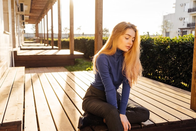 Piękna młoda blond dziewczyna w niebieskim swetrze siedzi na zewnątrz w słońcu, pozowanie