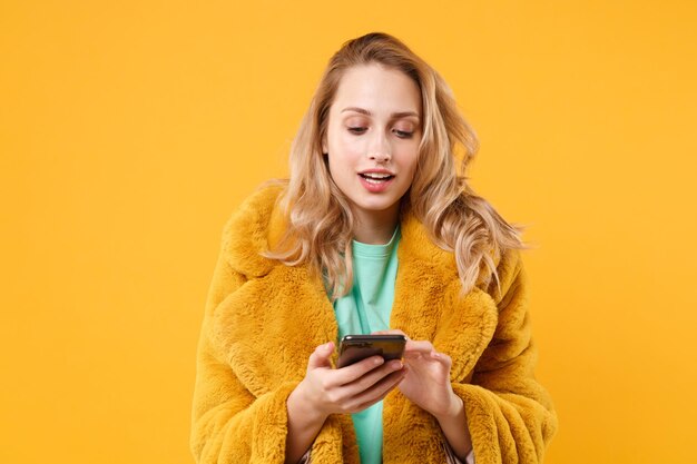 Piękna młoda blond kobieta dziewczyna w żółtym futrze pozowanie na białym tle na pomarańczowym tle portret studio. Koncepcja życia emocje ludzi. Makieta miejsca na kopię. Za pomocą telefonu komórkowego wpisując wiadomość sms.