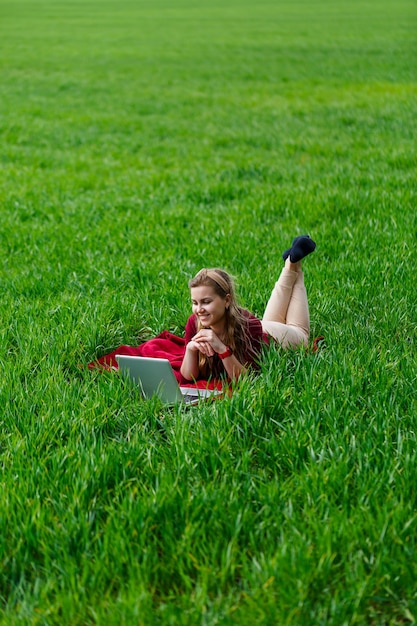 Piękna młoda blond kobieta leży na zielonej trawie w parku z laptopem i pracy. Błękitne niebo z chmurami. Dziewczyna uśmiecha się i cieszy dobry dzień. Pracuj nad naturą w słoneczny dzień.