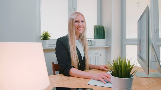 Piękna młoda blond kobieta siedzi w miejscu pracy z komputerem i trzyma za rękę mysz patrząc na...