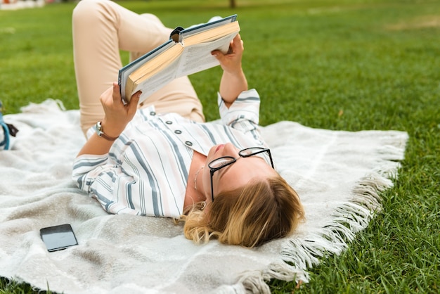 Piękna młoda blondynka relaksuje się na trawniku w parku, czytając książkę leżąc na plecach na kocu