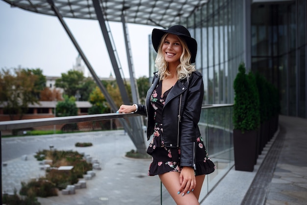Piękna młoda blondynka w skórzanej czarnej kurtce, ładnej krótkiej sukience i kapeluszu spaceruje po parku Zaryadye w Moskwie.