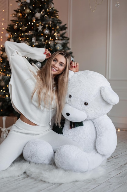 Piękna młoda blondynka z ładnym śnieżnobiałym uśmiechem, ubrana w modny biały strój z kapturem, siedzi obok misia w pobliżu choinki w domu