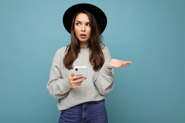 Piękna młoda brunetka kobieta myśli sobie czarny kapelusz i szary sweter trzymając smartfon patrząc z boku SMS-y na białym tle na tle