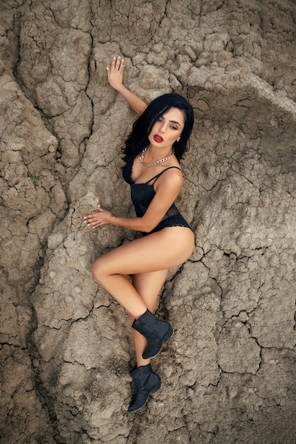 Piękna młoda brunetka w seksownym czarnym body na suchym piasku. Modelka o idealnej sylwetce pozuje w pustym kamieniołomie.