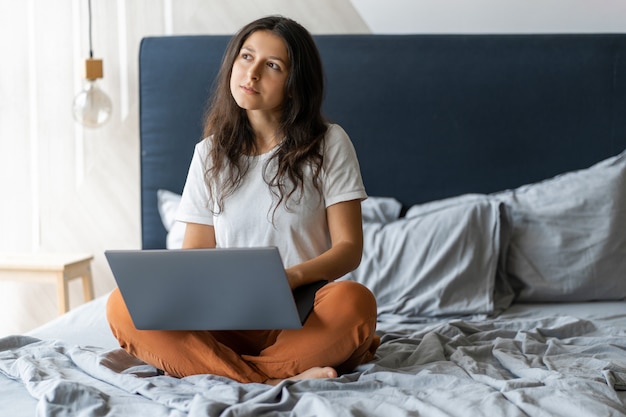 Piękna młoda brunetki dziewczyna z laptopu obsiadaniem na łóżku. Stylowe nowoczesne wnętrze. Przytulne miejsce pracy. Zakupy przez Internet