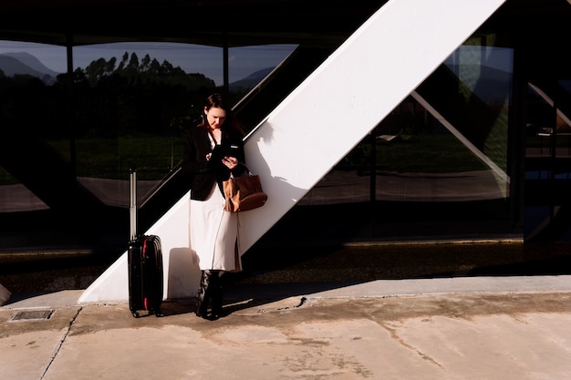 Zdjęcie piękna młoda dorosła kobieta w podróży służbowej, wzywając taksówkę na lotnisko lub sprawdzając pocztę e-mail w dzielnicy biznesowej lub w parku przemysłowym niosąc walizkę. pomysł na biznes