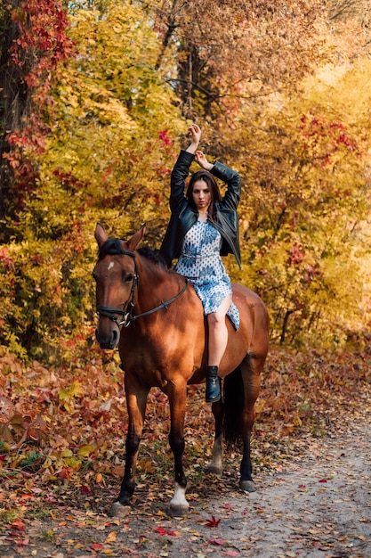Piękna młoda dziewczyna siedzi okrakiem na koniu w sukience i butach z podniesionymi rękami