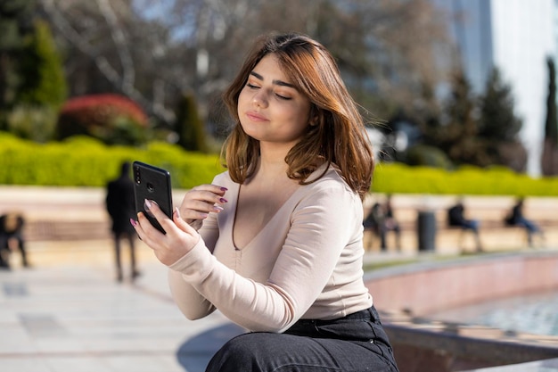 Piękna młoda dziewczyna siedzi w parku i patrzy na swój telefon Zdjęcie wysokiej jakości
