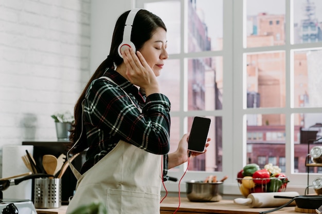 Piękna młoda dziewczyna słuchania muzyki w słuchawkach stojąc przy blacie kuchennym. azjatycka japońska gospodyni domowa korzystająca z telefonu komórkowego i słuchawek cieszyć się piosenkami podczas gotowania w domu w domu