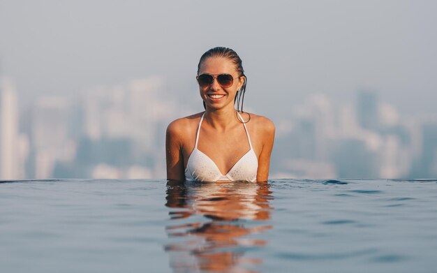 Piękna młoda dziewczyna w białym bikini pływająca i stojąca na krawędzi basenu na szczycie hotelu i patrząca na panoramę miasta