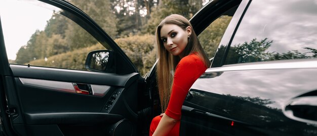 Piękna młoda dziewczyna w czerwonym kombinezonie siedzi za kierownicą czarnego samochodu na pustej drodze w lesie