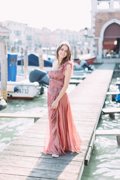 Piękna młoda dziewczyna w różowej sukience stoi na drewnianym moście między kanałem weneckim i ciesząc się miastem.