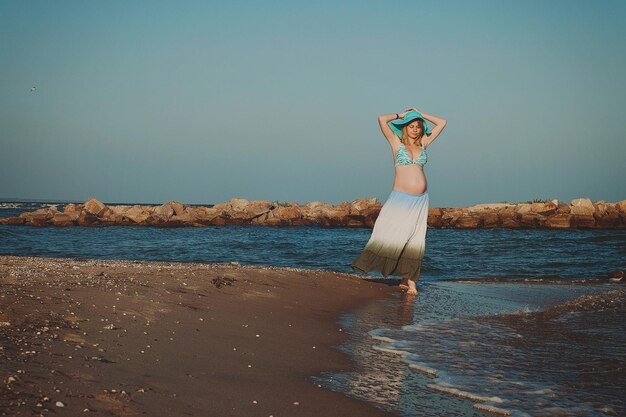 Piękna młoda elegancka blond kobieta w ciąży w niebieskim kapeluszu spacery po plaży, pobrzeże o zachodzie słońca.