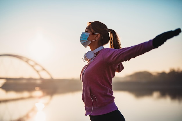 Zdjęcie piękna młoda i sprawna kobieta w dobrej kondycji bieganie i jogging samotnie na ulicy mostu miasta. nosiła maskę ochronną na twarz, aby chronić się przed infekcją wirusową lub alergiczną. zachód słońca w tle.
