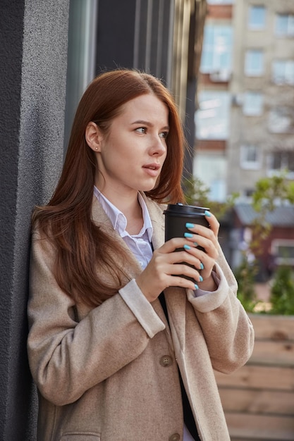 Piękna młoda kaukaska kobieta w stylowym beżowym płaszczu pijąca kawę na zewnątrz