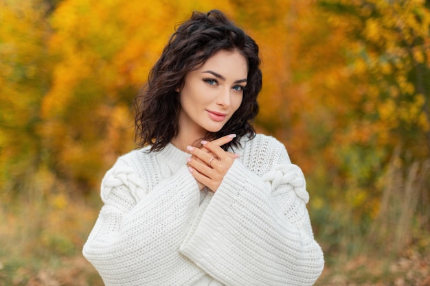 Piękna młoda kędzierzawa stylowa dziewczyna modelka z uroczą świeżą twarzą w modnym swetrze z dzianiny spaceruje w jesiennym złotym parku Kobiecy jesienny portret ładnej kobiety