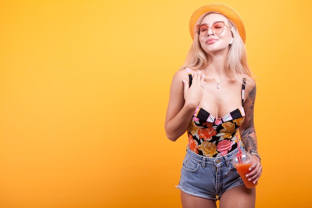 Piękna młoda kobieta ciesząca się świeżym sokiem pomarańczowym z kapeluszem i okularami przeciwsłonecznymi w studio